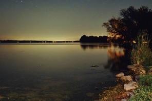 photo at night of lake shore with grassy bank, glowing horizon, and sky with bright stars