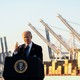 Joe Biden speaking from a lectern in a port, stacks of shipping containers visible on the background.