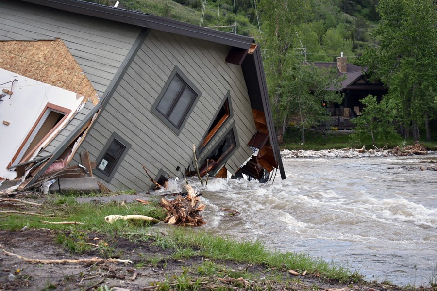 A house leans steeply into a flooding river that is undercutting it and dragging it down.
