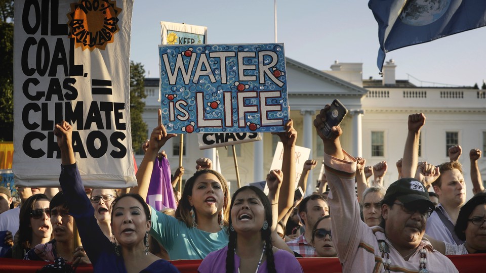 Supporters of the Standing Rock Sioux have signs reading “Oil, Coal, Gas = Climate Chaos,” and “Water is Life.” Their fists are raised, with the White House in the background.