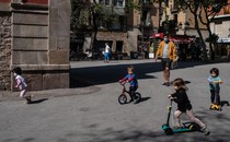 A man in a mask oversees a group of children playing on bikes and scooters in a plaza outside.