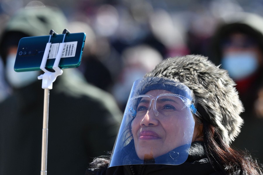 A woman wearing a clear face shield holds up a smart phone on a selfie stick.