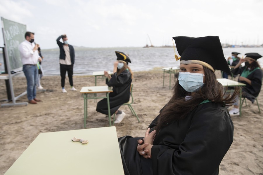 Young students wearing face masks and graduation caps and gowns sit at widely spaced desks set up on a beach.