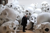 A person stands in an exhibit space amid a large pile of giant human-skull sculptures.