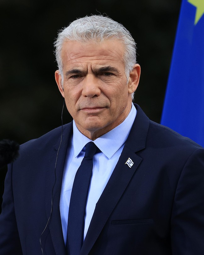 Yair Lapid, wearing a navy suit and tie, stands in front of a pair of microphones, with the EU flag in the background.