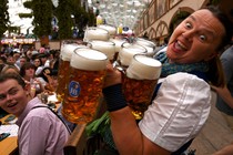 A person smiles while carrying ten large beer mugs in a beer hall.