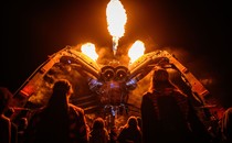 Fire erupts from the top of a large robotic spider sculpture as people look on below.