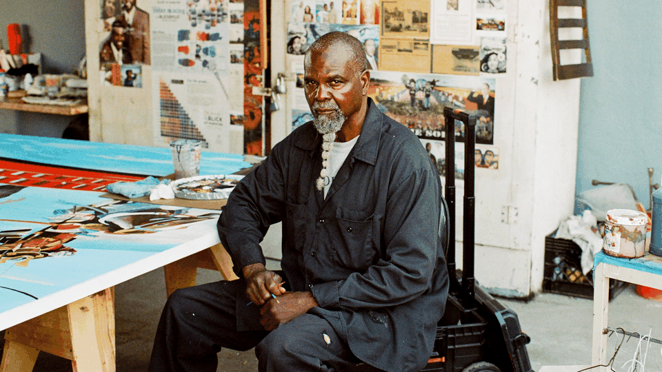 MR WASH sits in a chair facing the camera, with a solemn expression. The room around him is covered in artwork.