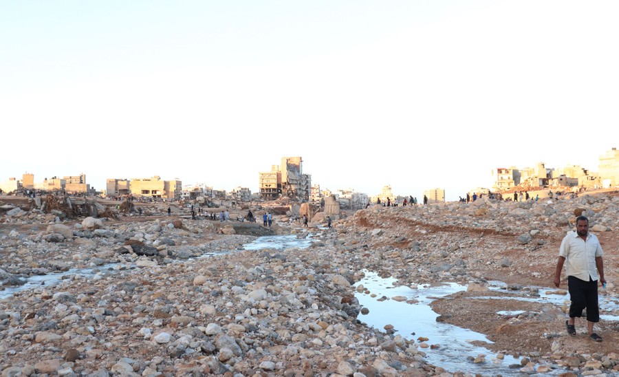 People walk over loose rocks, mud, and debris, among many damaged buildings.