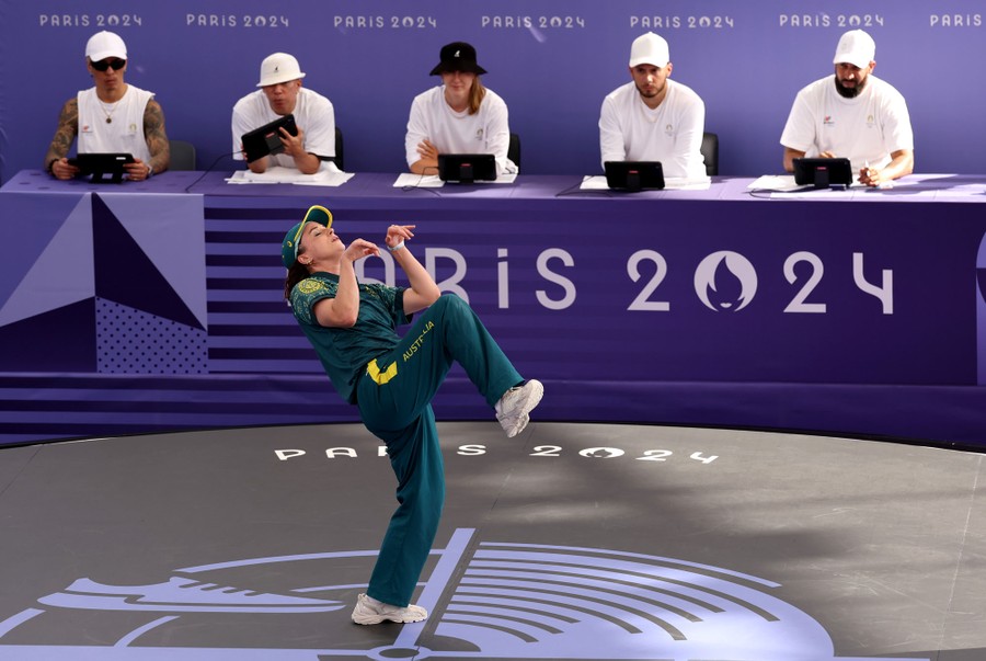 A breakdancer performs for a panel of judges at the Olympics, photographed in an unusual pose resembling a hopping kangaroo.