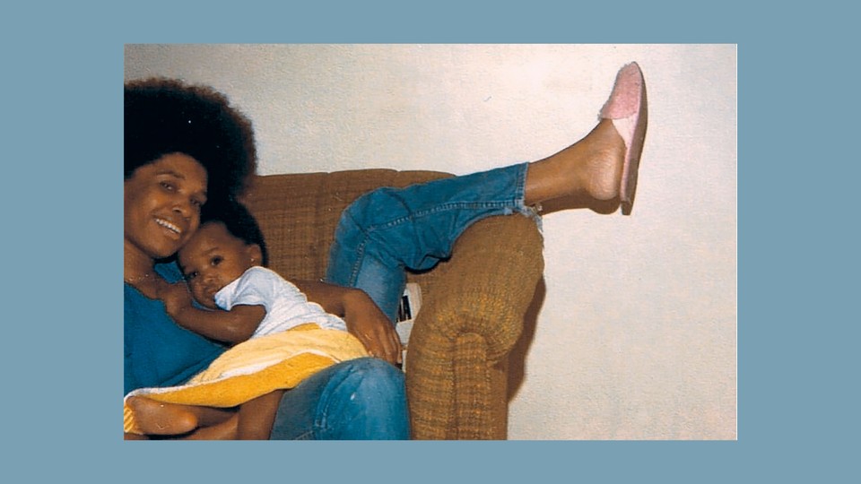 A woman sitting in a brown chair with her leg thrown over the arm holds a baby while looking at the camera.