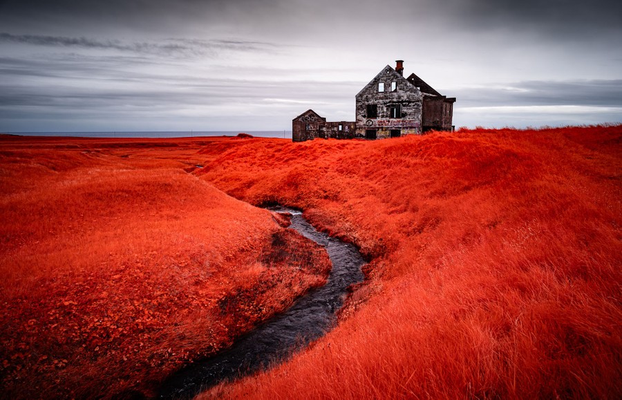 A dilapidated and abandoned stone building stands in a rolling field, with the surrounding grass appearing orange in color.