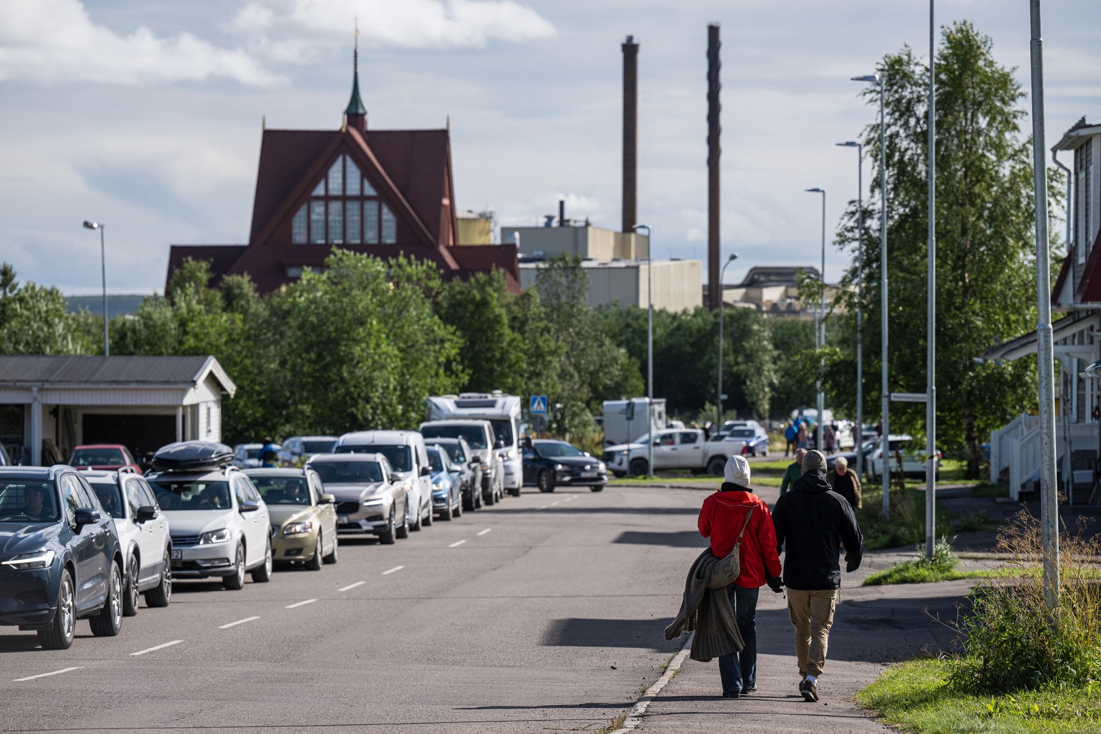 People walk along a road, with the top of a wooden church visible in the distance.