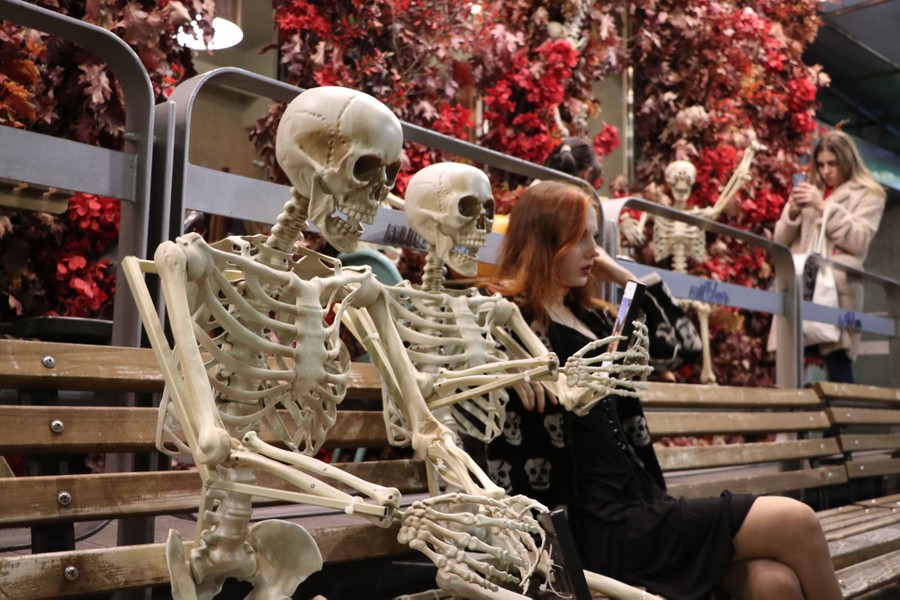 A person sits on a bench beside two decorative skeletons.