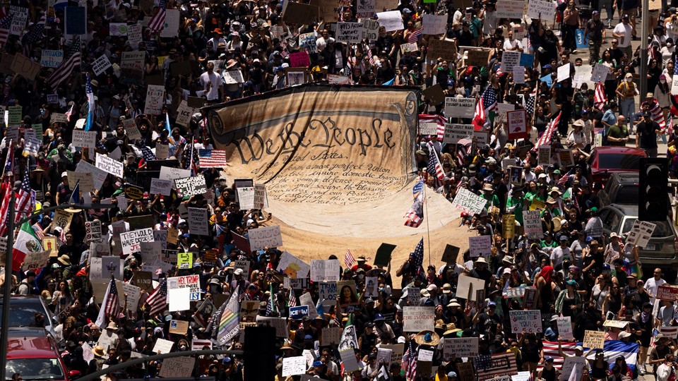 A large crowd of protestors at a previous "No Kings" rally holding up signs and surrounding a large replica of the constitution.