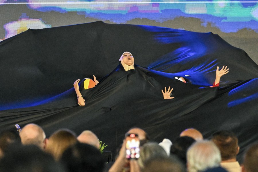 Performers dance beneath a large sheet of black fabric with several holes cut in it, poking hands and a head above.