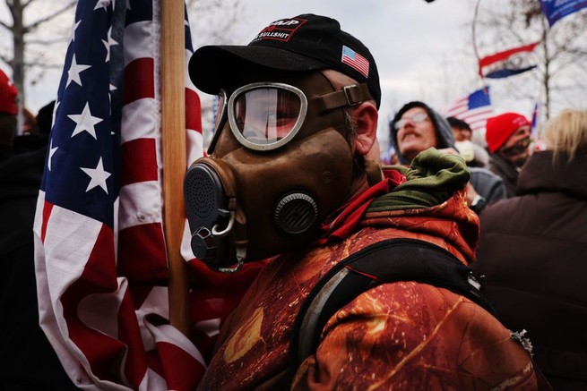 Man in gas mask at the capital on January 6, 2021, holding an American flag and wearing a Trump hat