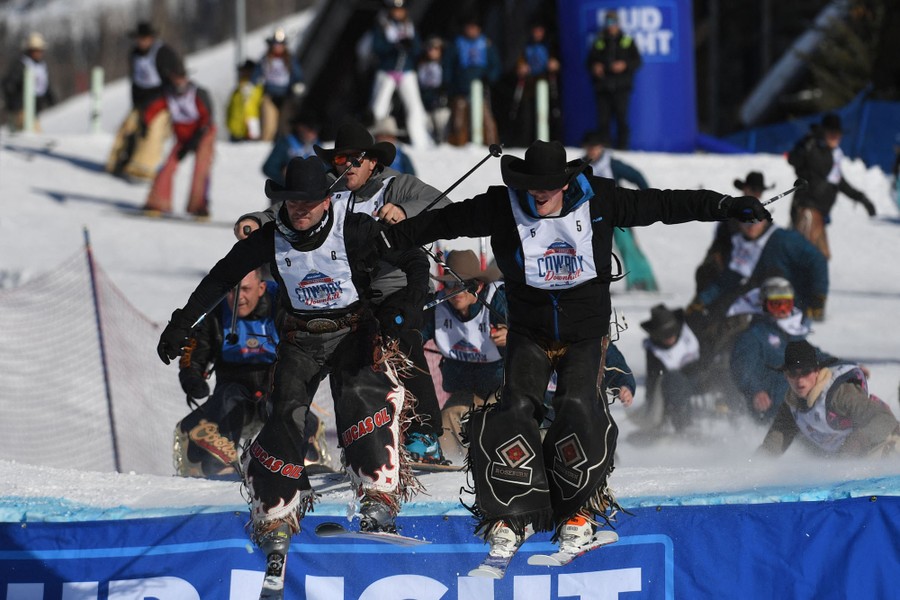 People wearing cowboy hat, chaps, and racing bibs compete in a downhill ski race.