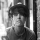 A black and white photo of a person with dark hair wearing a narrow hat and a thin sweater beside a brick wall.