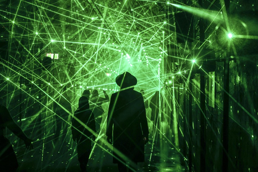 People walk through a hall of mirrors lit only by many reflected green laser beams.