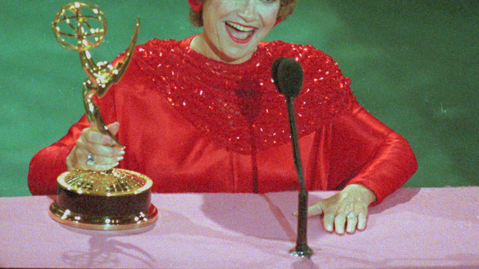 Woman in a red dress holding a golden Emmy trophy, smiling in front of a microphone.