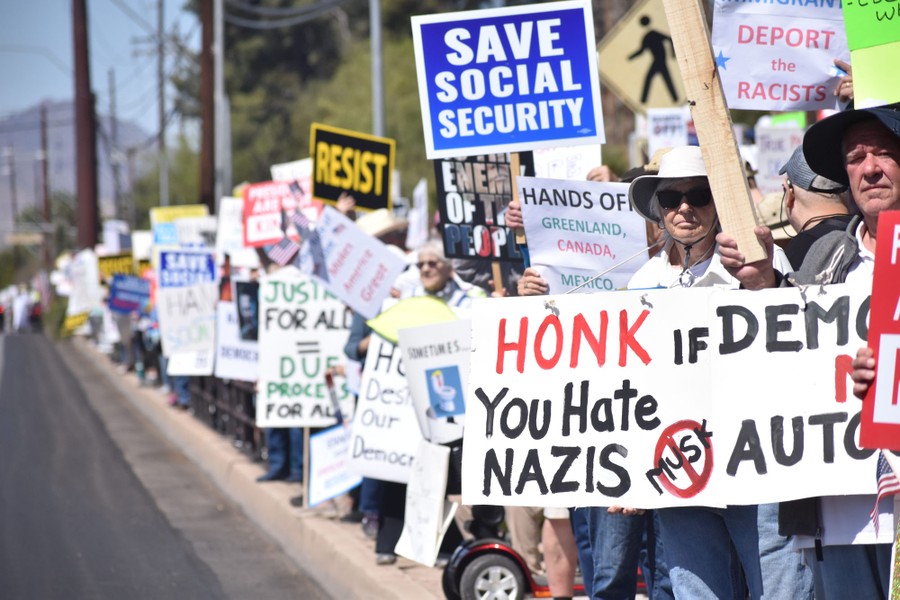 A long line of protesters stands on the curb of a street, holding many signs, including one that reads "Save Social Security."