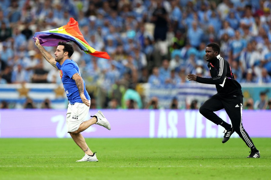 A security guard chases a man carrying a rainbow flag on a soccer pitch.