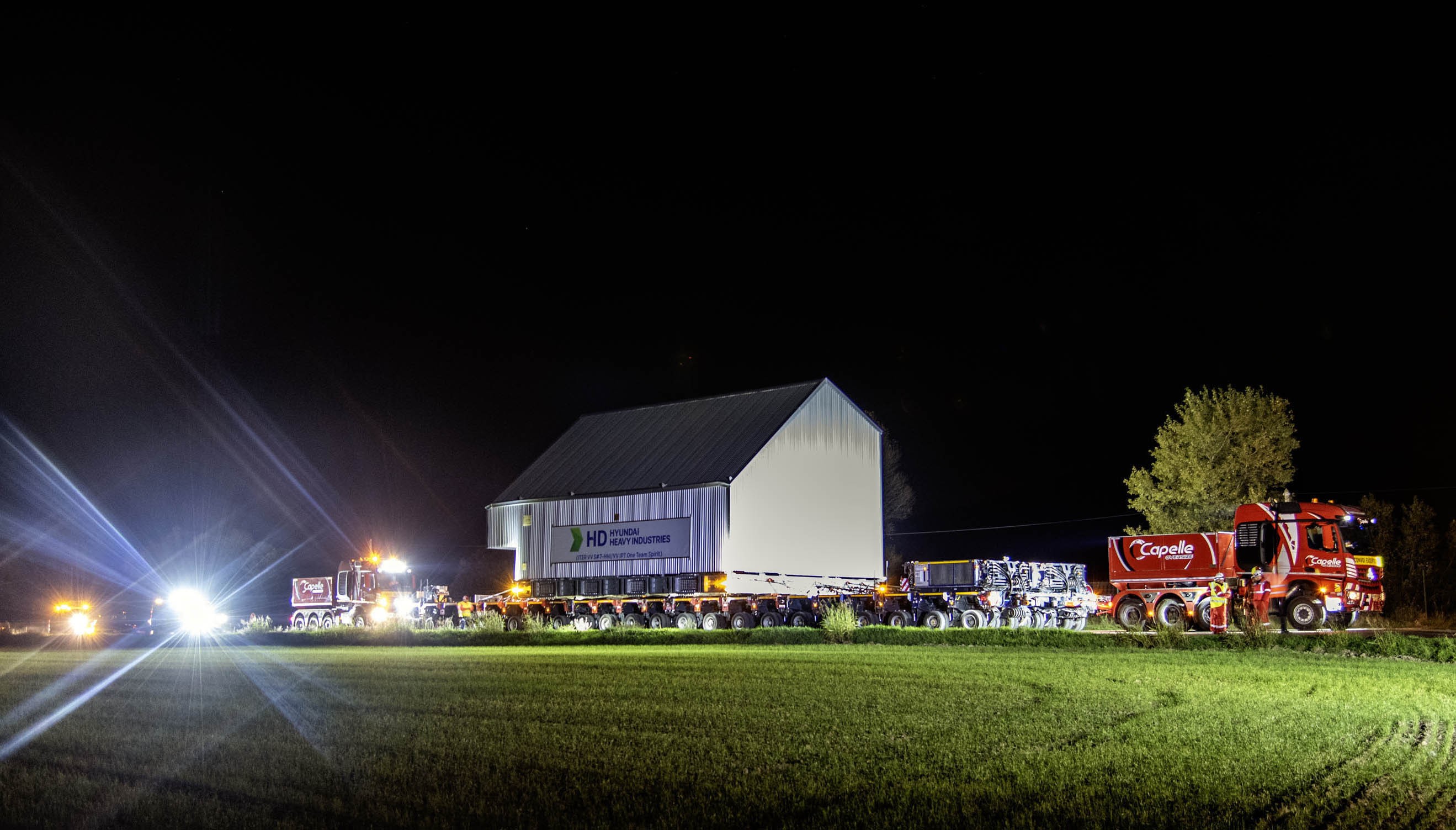 A truck pulls a long trailer with a large metal cabin-like shipping container on it