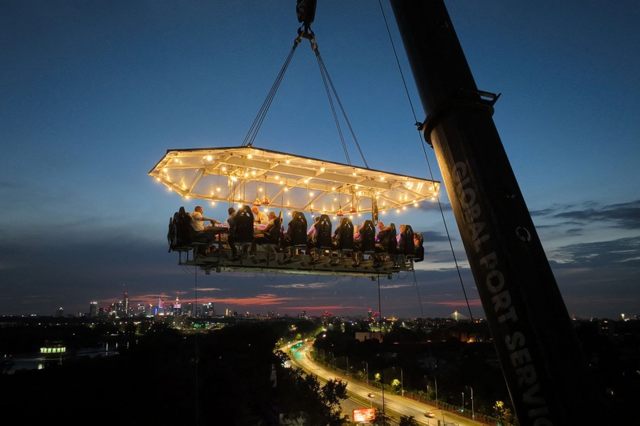 About 20 people are suspended in chairs around a specialized dinner table that is held high in the air by a tall crane.