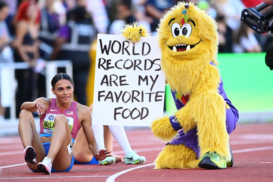A furry mascot kneels on a track next to an athlete after a race, holding a handmade sign that reads "World records are my favorite food".