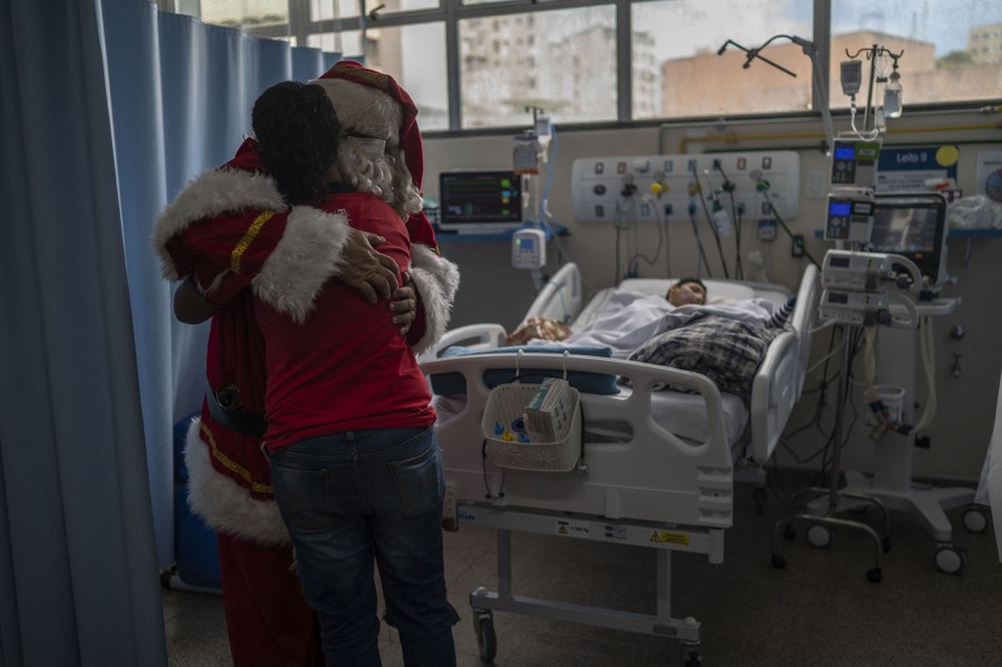 A man dressed as Santa Claus hugs the mother of a child who lies in a hospital bed.