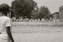 Photograph of an African American boy watching a group of people, some carrying American flags, marcing past to protest the admission of the "Little Rock Nine" to Central High School