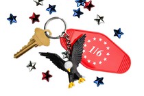 On a white background, a collection of scattered 3-D confetti-style stars are strewn about. They are red, white, and blue. In the middle is a keychain with a single gold key, a red tag reading "1/6" with a ring of white stars printed on it, and a miniature bald eagle.