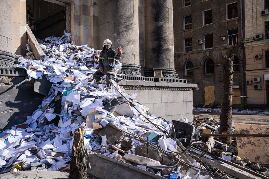 A firefighter stands atop a pile of papers and building debris, near an entrance to a damaged building.