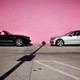 An image of a black convertible (left) and a silver sedan (right) that are parallel-parked along a pink wall, facing each other