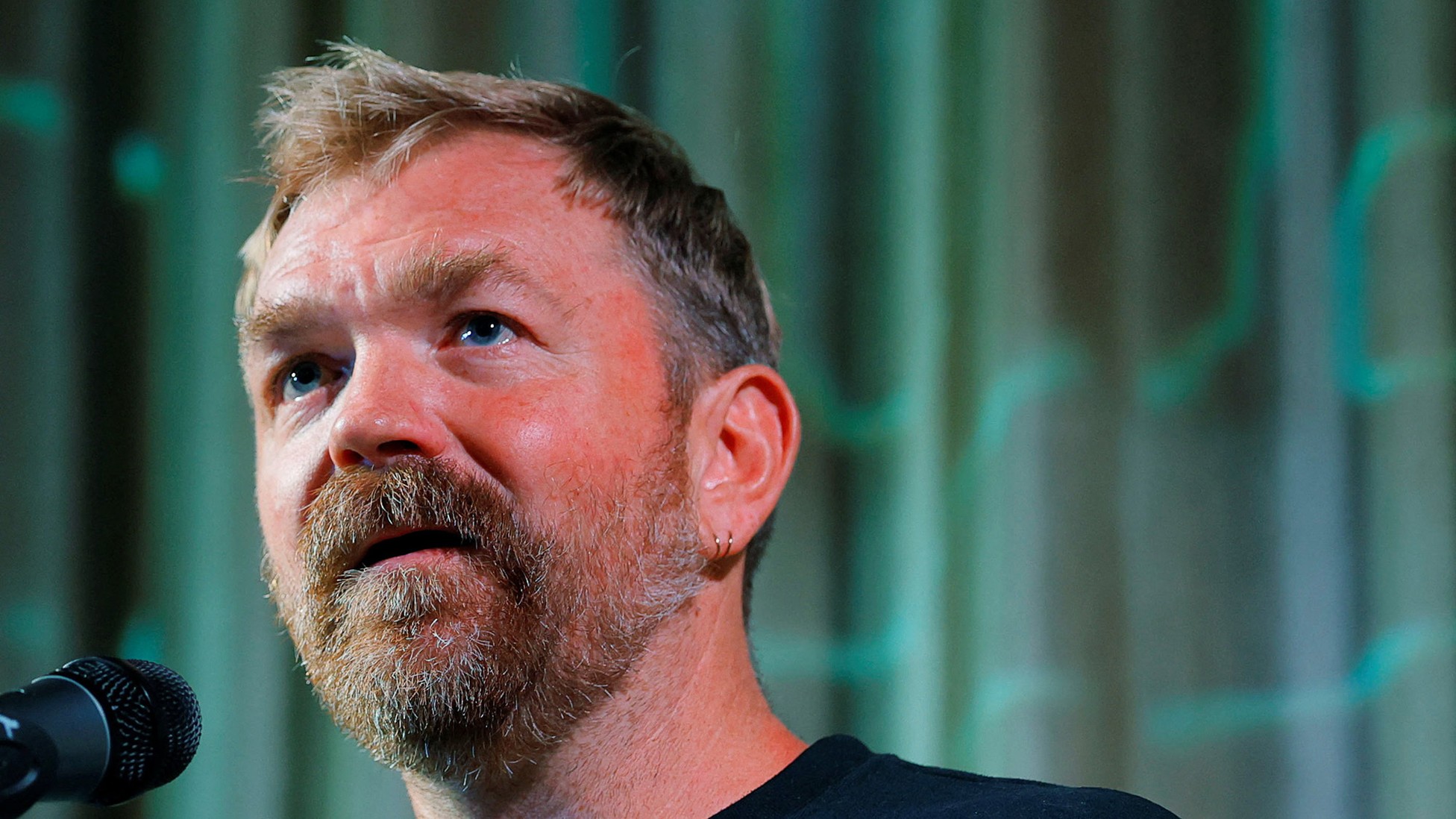 عکسی از گراهام پلاتنر در حال سخنرانی با میکروفون در پسزمینهای آبی-سبز