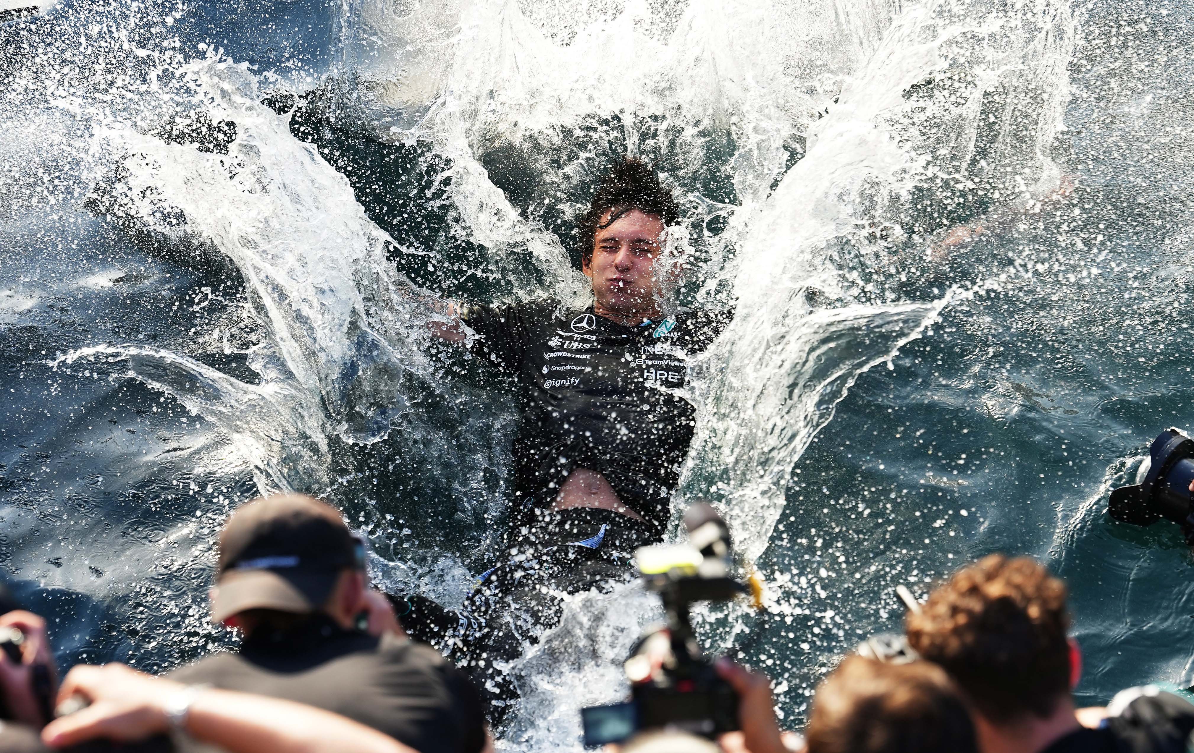 A race car driver makes a splash after jumping into a harbor in celebration, as others take photographs.