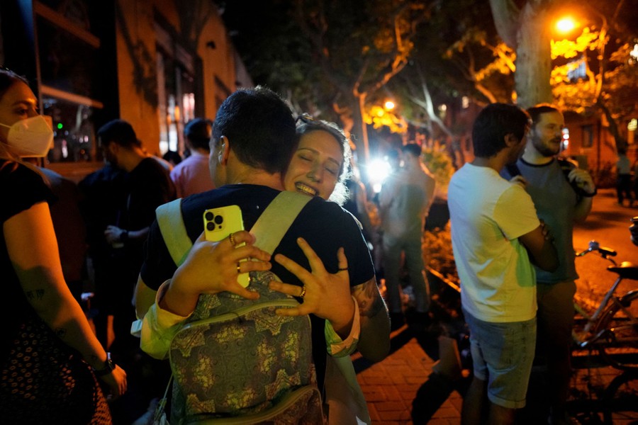 People gather on a city street at night, talking and embracing.