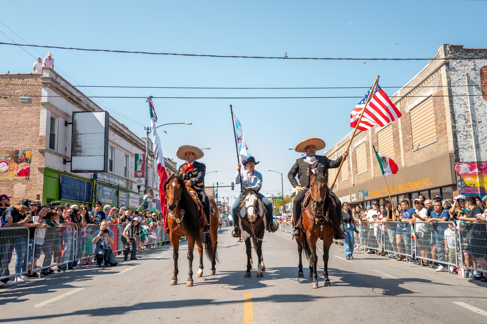 Three people on horseback, two wearing mariachi costumes, carry American and Mexican flags on a parade route.