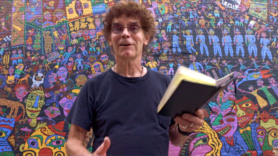 A curly haired artist reads from a book, in front of a mural he painted.