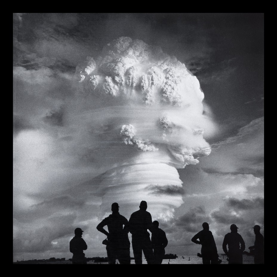 black-and-white photo of silhouetted group of soldiers standing on shore with enormous swirling mushroom cloud in distance
