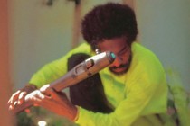 André 3000 crouching in a neon yellow-green shirt holding a flute