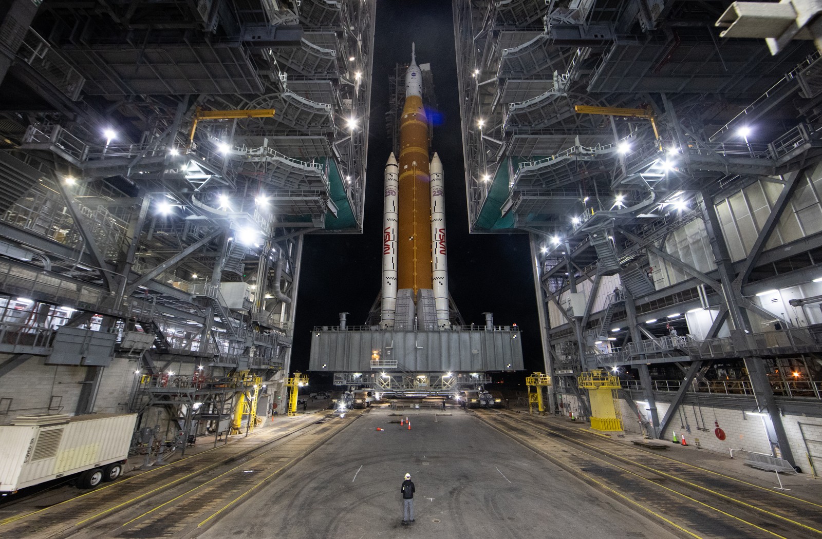 A tall rocket is rolled out of a launch facility building on top of a large tracked vehicle.