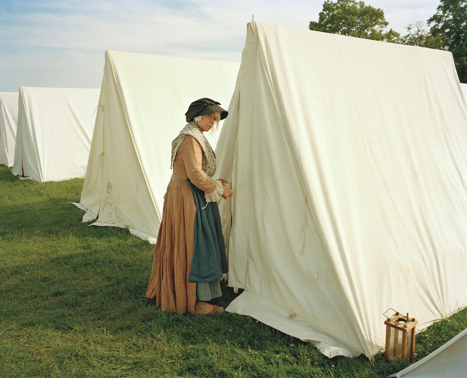 photo of woman in colonial dress, apron, and hat looking sideways at viewer as she enters one of a row of white-cloth triangular tents pitched on the grass at Gloucester, MA during a reenactment of the Battle of Bunker Hill