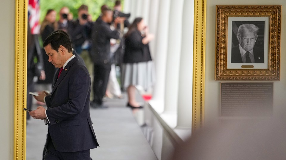 Marco Rubio tapping at his phone while walking past Donald Trump's portrait