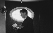 Black-and-white photograph of John Hollander reading from loose folded pages