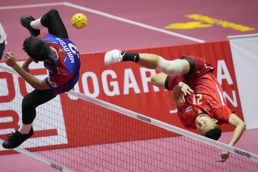 Two athletes are seen in mid-leap, twisting to kick a ball over a net.