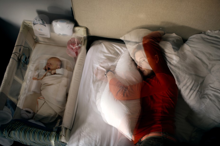 Photo illustration: from overhead, mother hugging pillow on bed next to swaddled baby in bassinette