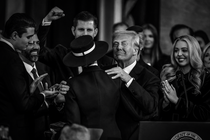 A black-and-white photo of Donald Trump facing the camera in a suit with a slight smile, while embracing Melania Trump, whose back is toward the camera. They are surrounded by people who are clapping and smiling.
