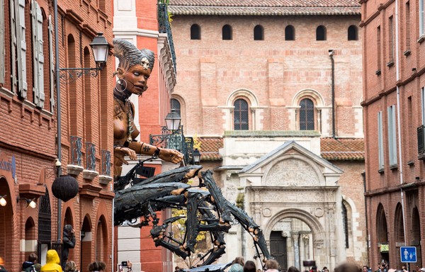 A giant mechanical puppet of a mythical human-scorpion creature moves through a narrow street.